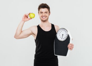 Photo of smiling young sportsman holding scales and apple isolated over white background. Looking at camera.
