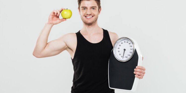 Photo of smiling young sportsman holding scales and apple isolated over white background. Looking at camera.